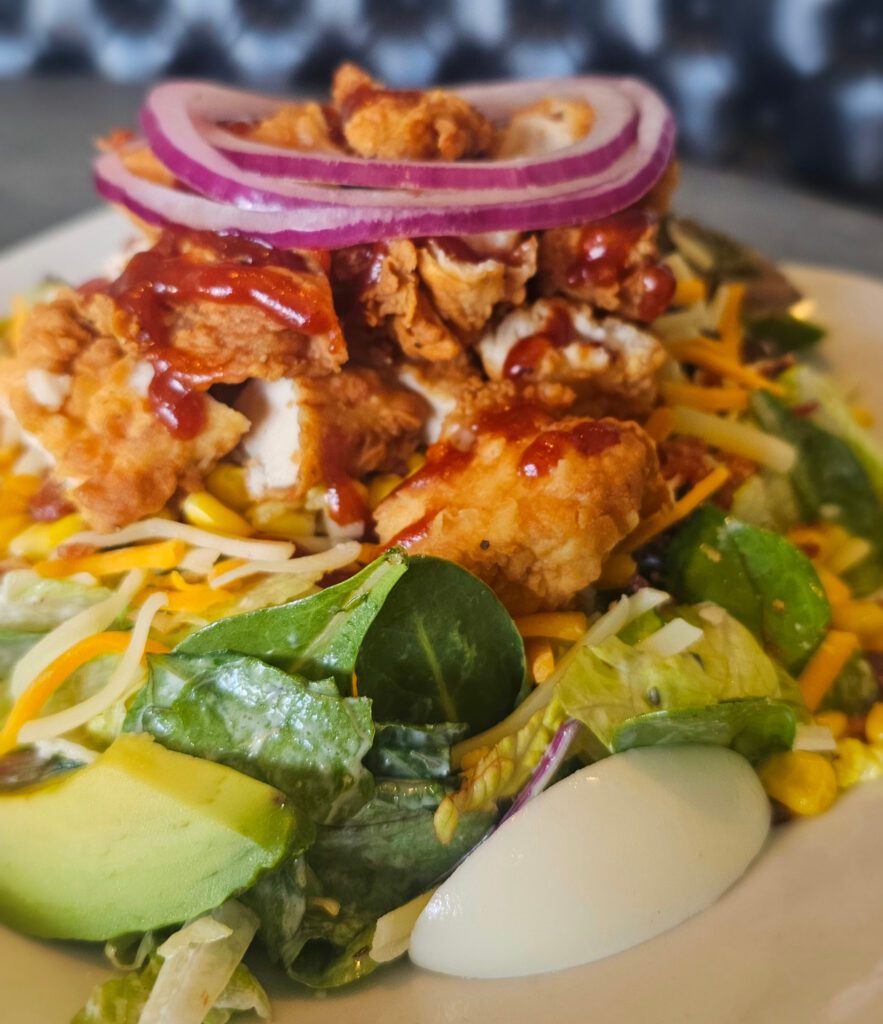 California Cobb: Mixed greens, romaine & croutons tossed in buttermilk ranch dressing with bacon, hardboiled egg, roasted corn, avocado, cheddar/jack cheese, red onion; topped with crispy chicken tenders with a honey barbecue drizzle.